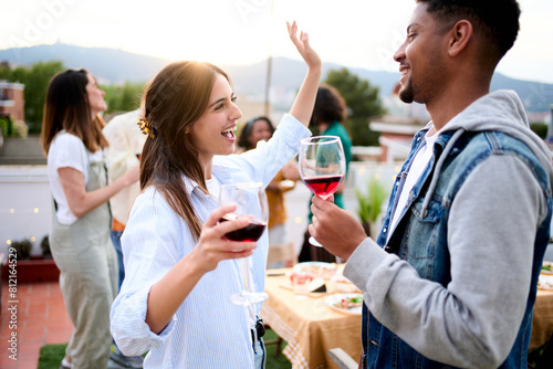 Excited multiracial young couple celebrating rooftop party drinking red wine. Friends dancing at sunset date weekend gathering. Cheerful gen z people enjoying romantic leisure outdoors.