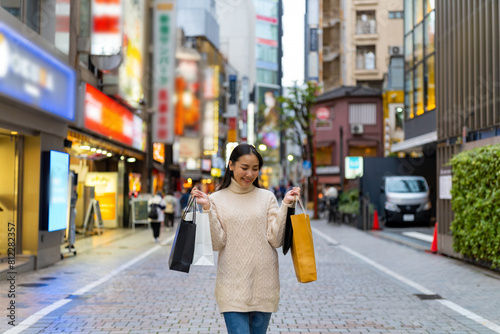 Happy Asian woman holding shopping bag walking at Shibuya district, Tokyo, Japan in evening. Attractive girl enjoy and fun outdoor lifestyle travel urban city street and shopping on holiday vacation.