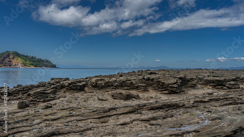 The seabed was exposed at low tide. Attached mollusks are visible on the layered stones. Puddles of water. White boats in the ocean. A hill against a blue sky and clouds in the distance. Madagascar.
