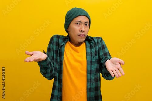 Young Asian man, dressed in a beanie hat and casual shirt, is making an I don't know Italian gesture, with open arms, raised shoulders, and showing both palms while standing against yellow background.