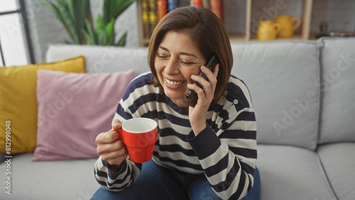 Smiling middle-aged hispanic woman talking on phone while holding a red mug indoors
