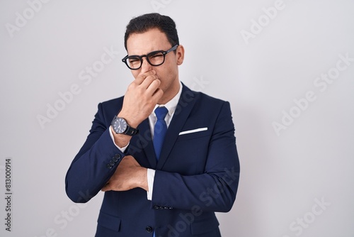 Young hispanic man wearing suit and tie smelling something stinky and disgusting, intolerable smell, holding breath with fingers on nose. bad smell