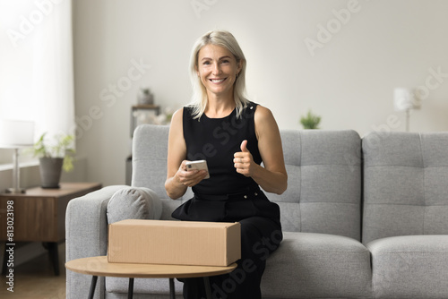 Happy satisfied mature Internet shop customer showing like sign, thumb up hand gesture, looking at camera, smiling, holding mobile phone, sitting at paper cox, cardboard package at home