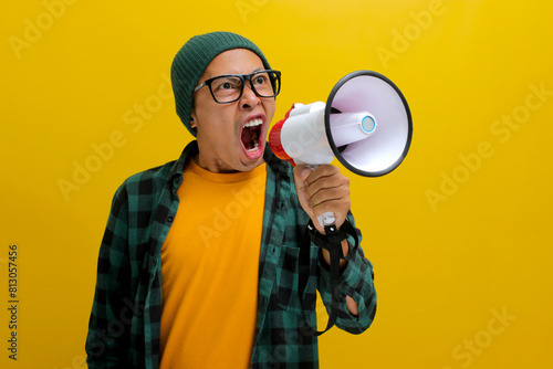 Young Asian man in a beanie hat and casual shirt shouts through a megaphone, expressing annoyance and frustration with angry yelling. Isolated on a yellow background