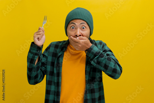 Surprised young Asian man, clad in a beanie hat and casual shirt, covers his mouth in shock and embarrassment, displaying a shocked expression after making a mistake, while holding a fork