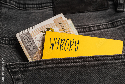  Yellow card with a handwritten inscription "Wybory", inserted into the pocket of gray pants jeasnow, next to Polish banknotes PLN (selective focus), translation: elections