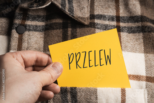  Yellow card with a handwritten inscription "Przelew", a shaft in the hand, protruding from a brown plaid shirt (selective focus), translation: transfer