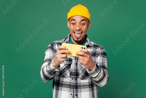 Young man wear shirt blue t-shirt yellow hat use play racing app on mobile cell phone hold gadget smartphone for pc video games isolated on plain green background studio portrait. Lifestyle concept