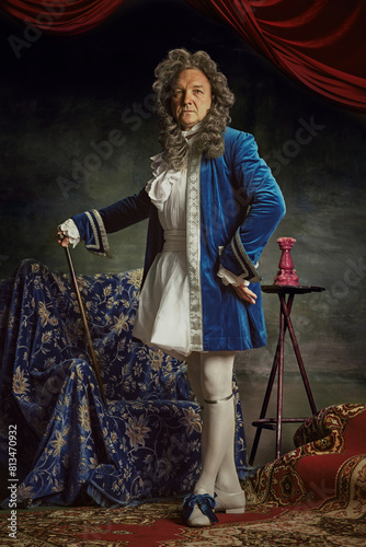 Full-length portrait of elderly man dressed as medieval person, aristocrat with confident expression against vintage studio background. Concept of comparisons of eras, fusion of modernity and history.