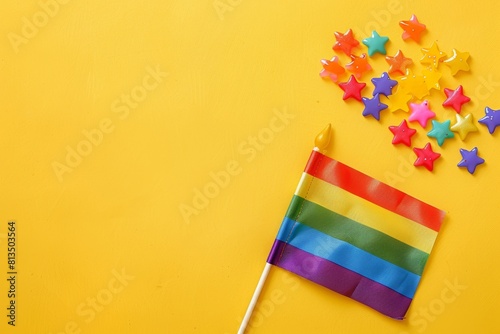 The flag on a stick and rainbow candy stars lay flat on a yellow background with copy space, in the concept of an LGBT flag. Pride Day themed rainbow and star toothpicks with pride flags, top view