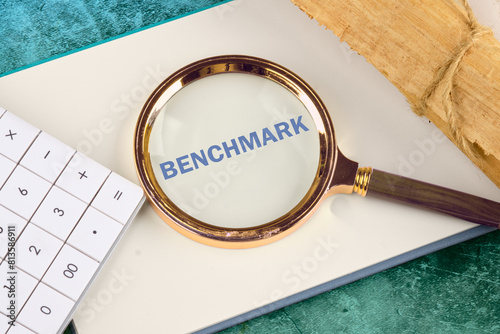 Business concept of benchmark. A word BENCHMARK written through a magnifying glass on a blank sheet of notepad next to a calculator and a roll of papyrus