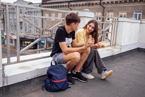 Two teenagers are sitting on the parapet on the roof of the house and eating Asian food