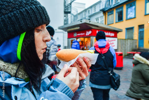 Reykjavik, Iceland - march 15th, 2023: Close up caucasian woman eat traditional tasty hotdog by famous red kiosk stand in downtown Reykjavik.Famous traditional icelandic foods