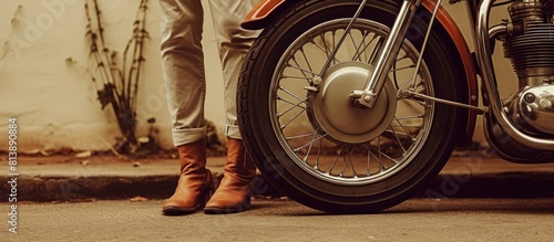 A person wearing vintage brown quadrille pants and brown sneakers stands next to an old motorcycle showing their legs and feet in the copy space image