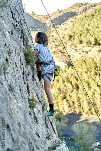 A boy is scaling a steep cliff, clinging to the rock wall as she practices climbing in extreme mountain terrain