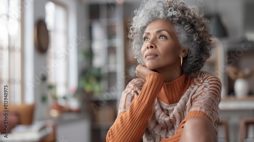 A thoughtful senior biracial woman indoors, gazing upwards with a hand on her chin, in a cozy home setting.