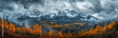 A panoramic view of the Rocky Mountains, showing larch trees in autumn colors with snow on top and dark storm clouds overhead. 