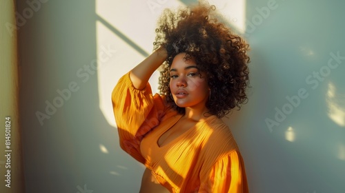 a Plus size woman with curly hair, wearing an orange blouse, standing against a wall