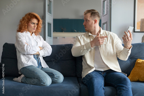 Spouses having conflict, angry man shouting at wife, pointing at her mobile phone. Defensive husband bringing arguments, explanations