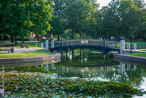 Green trees and pond with reflective surface. Pedestrian bridge over clear smooth pond. Central College campus in Pella, Iowa. 