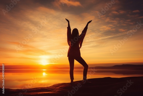 Woman's silhouette stands with arms raised against a vibrant sunset over calm waters