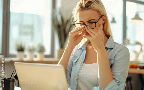 Stressed woman massaging nose bridge, holding glasses, laptop in front.