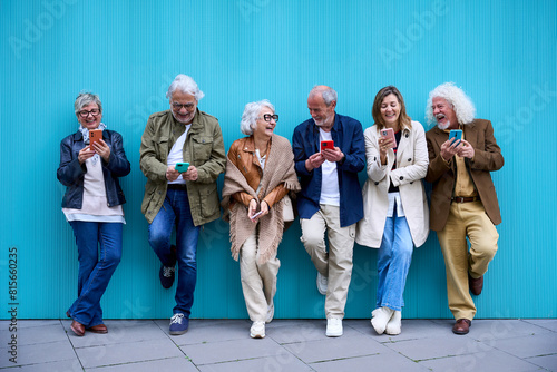 Group of elderly Caucasian laughing together using cell phone standing leaning on blue wall. Happy old friends with gray hair gathered having fun looking mobile outdoor. Mature people enjoying devices