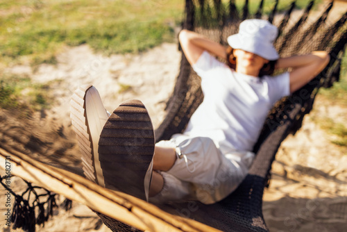 A young woman in casual clothes has her hands behind her head and is lying in a hammock. A girl in a panama hat relaxes and dozes in nature.