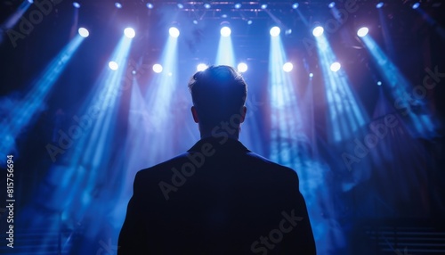 The man standing on the stage is looking at the audience. He is illuminated by a spotlight. He is wearing a dark suit. The background is dark.