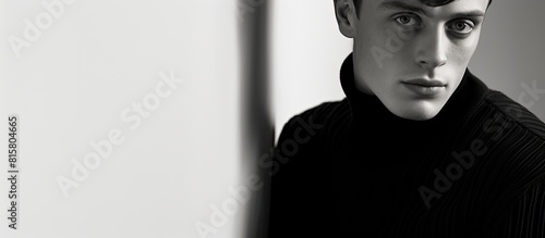 A melancholic young man dressed in a black turtleneck leans against a white wall and gazes serenely at the camera in a black and white photograph The image portrays a psychological profile with avail