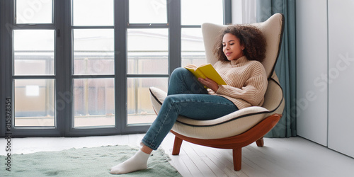 Young woman in casual clothes, reading a book in a comfortable armchair at home