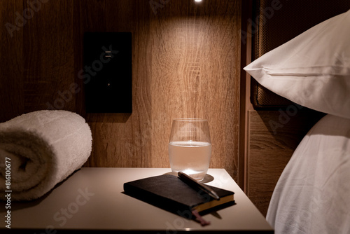 Hotel room table illuminated by small auxiliary lamp with agenda, pen glass of water and rolled towel