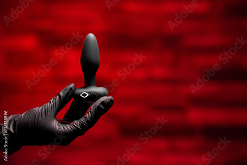 Silicone anal plug in a female hand in black latex gloves on a black background. Massager with multiple speeds. Sex shop products, adult gifts for couples.