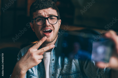 Excited male blogger showing peace fingers and taking funny pictures on mobile camera indoors, young emotional hipster guy making photos for social networks connected to wifi on smartphone device
