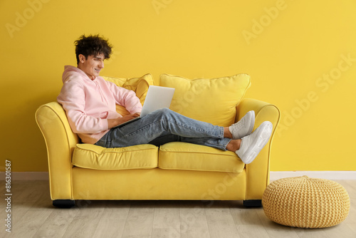 Young man resting on soft sofa with laptop near yellow wall