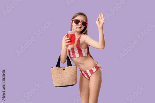 Cute little girl in swimsuit with beach bag and glass of lemonade waving hand on purple background