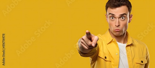 A young man with an expression of embarrassment on his face is pointing towards something on a yellow background in a copy space image
