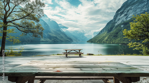 Serene mountain lake with wooden picnic tables, surrounded by lush green trees and towering mountains under a partly cloudy sky.