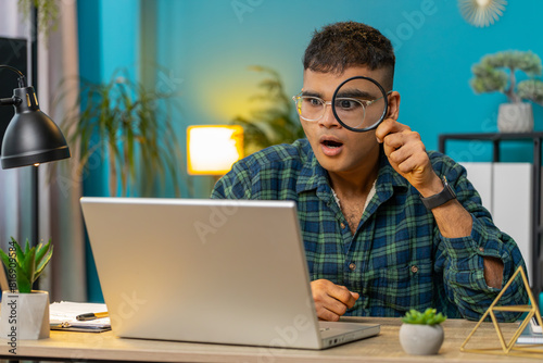 Surprised investigator researcher Indian businessman working at home office holding magnifying glass looking at laptop screen with wow expression, searching, analyzing. Amazed shocked freelancer guy