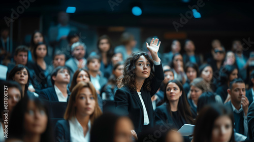 A young woman raises her hand in a crowded lecture hall filled with professional individuals, suggesting participation or inquiry in a formal setting.