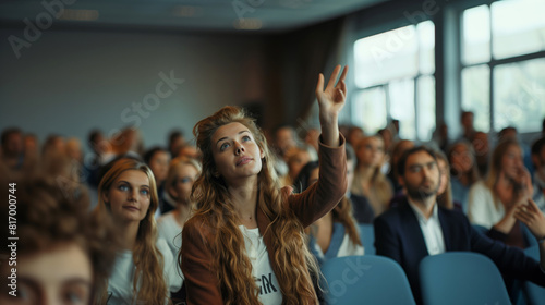 A woman raising her hand in a classroom or seminar setting, with attentive audience members in the background. The atmosphere suggests active participation and engagement.