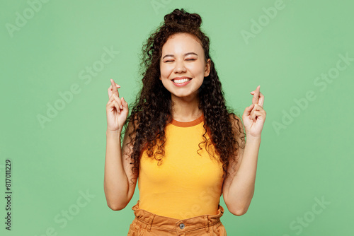 Young woman of African American ethnicity wear yellow tank shirt top waiting for special moment, keeping fingers crossed, making wish, eyes closed isolated on plain green background Lifestyle concept
