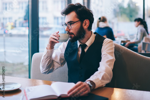 Fashionable hipster guy drinking hot coffee and looking at cafeteria window after reading interesting best seller, caucasian man pondering on literature during enjoying caffeine beverage indoors