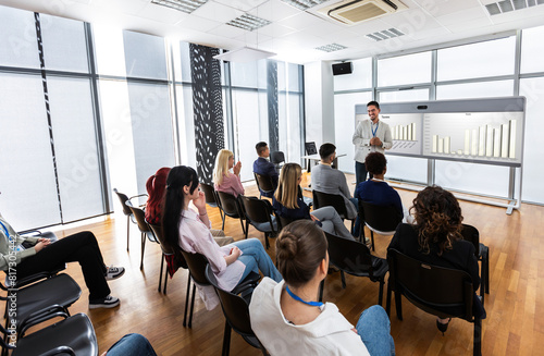 Male CEO standing in front of a multiracial group of colleagues. Multimedia classroom with smart board and graphics on them.