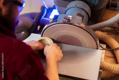 Artisan sanding wooden object on disc sander in assembling shop. Diy hobby enjoyer working in joinery, guiding wood against grinding machine rotating disc to create art