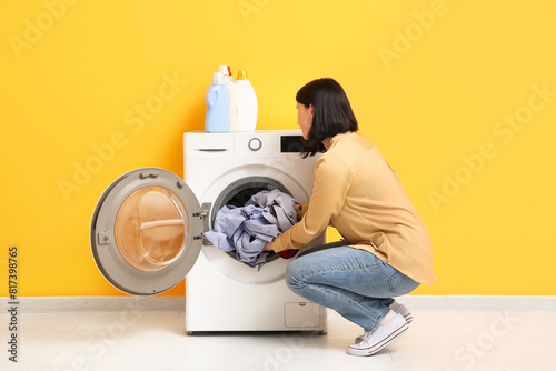 Young woman putting dirty clothes into washing machine on orange background