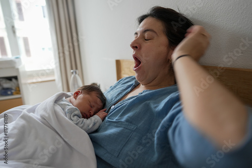 A mother yawns while holding her sleeping newborn baby on her chest, both in bed. The scene captures the tender and exhausting moments of early parenthood, illuminated by soft natural light.