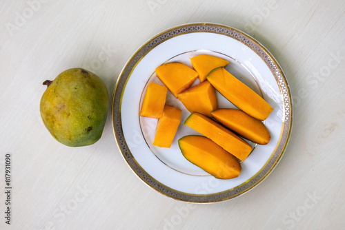 Peeled mango slices on a white plate on wooden background, a whole ripe mango placed beside. Top view