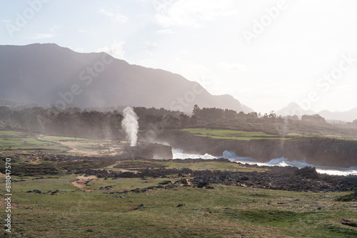A mountain range is in the background with a large steam coming out of the ground. The steam is white and billowing, creating a sense of mystery and wonder. The landscape is vast and open