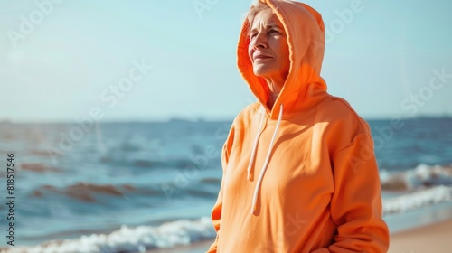 A woman in an orange hoodie is standing on the beach, looking out at the ocean. She is lost in thought, contemplating her life. AIG51A.